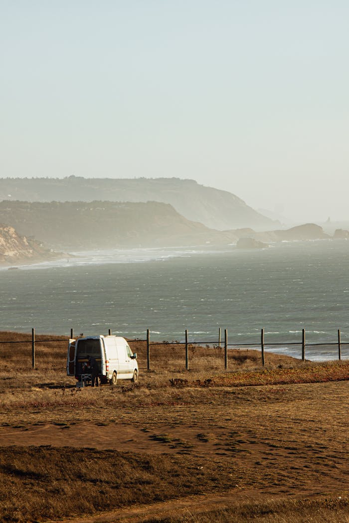 White camper van parked on a cliff overlooking the ocean with distant mountains, capturing a serene coastal landscape.